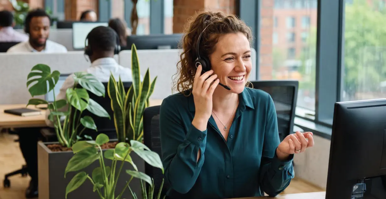 Smiling customer advisor wearing a telephone headset seated at a desk in a modern office environment.
